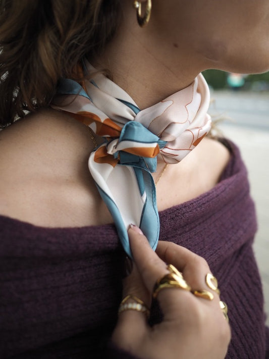Klassisk satintørklæde med smuk orange pæon på blå og cremefarvet baggrund
Ruby er et satintørklæde med et enkelt og elegant blomstermotiv af en orange, udfoldet pæon med smukke petroleumsblå blade. Baggrunden er cremefarvet med en blå konstrastkant, der giver det satintørklædet eksklusivt look.
Satinen tilfører en diskret glans og falder smukt, når tørklædet bindes.
Brug tørklædet
Om halsen som et klassisk silketørklæde
Om hoften i ægte Y2K-stil